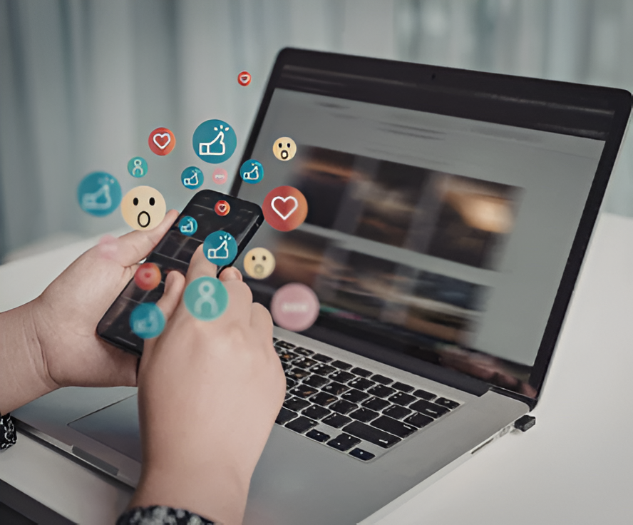 A person using a smartphone in front of a laptop, with colorful social media icons such as likes, hearts, and emojis appearing above the phone, symbolizing online interaction and social engagement.